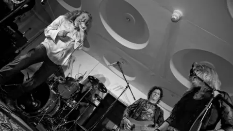 Richard Shakespeare A black and white image shows a live band in mid performance. On the left a man with long dark blond curly hair sings into a microphone, wearing a white shirt and dark trousers. Behind him, sits a drummer behind a drum kit, with a man with dark curly hair playing a bass guitar next to them. Further to the right, another man with long blond hair and a dark leather jacket sings into a microphone.