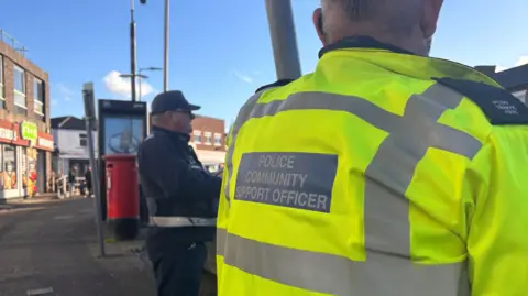 Cambridgeshire Constabulary A community support officer with his back to the camera. He wears a high-visibility coat and is stood outside during the day. 