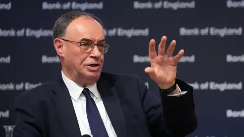 PA Media Governor of the Bank of England Andrew Bailey sits on a stage, addressing the audience and gesturing with an open hand. He wears a dark suit, white shirt, dark tie and glasses.