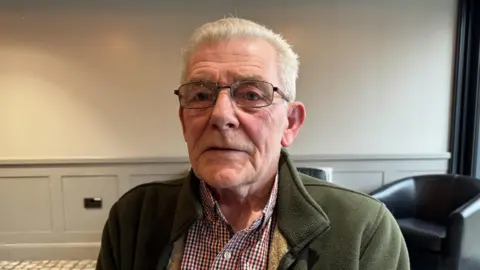 A white man with short grey hair and a green fleece sitting down  