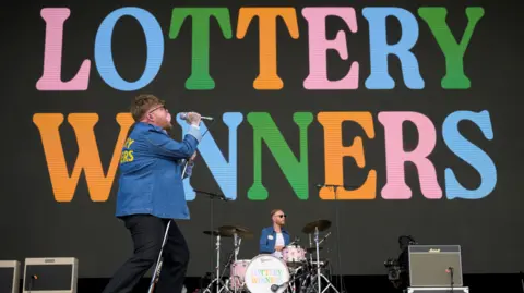 Getty Images The Lottery Winners on stage with the name of the band in colourful letters on a screen behind them.
