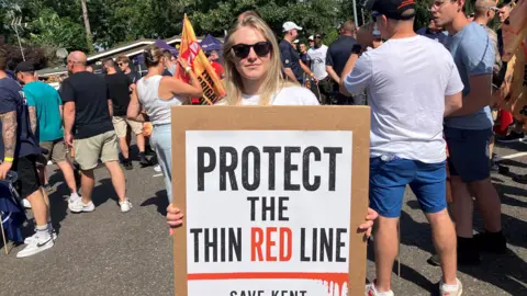 LDRS A blonde woman stands in a crowd holding a sign which reads 'protect the thin red line'