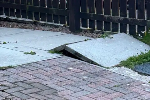 Simon Woodward A paving slab which has raised slightly with a fence in the background