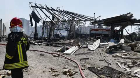 An emergency service worker dressed in a hard hat and reflective jacket looks on at the devastated petrol station after the explosion. The building is completely gutted by fire and is badly burnt. A fire engine can be seen nearby