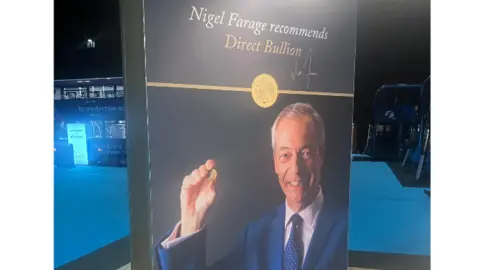  "Nigel Farage recommends Direct Bullion". Farage is wearing a blue suit with a pale pink shirt and a blue tie. He is smiling and holding up  a gold coin. 