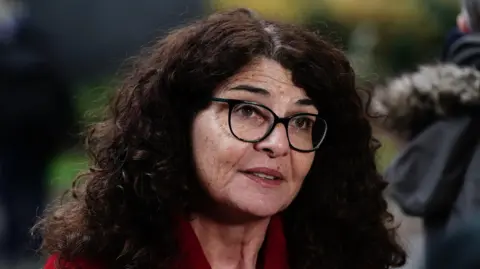 A woman with long curly hair wears glasses and a red top. Blurred in the background are some trees. 