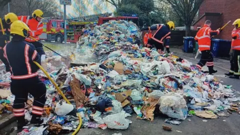 Cambridge City Council Firefighters are seen dousing down piles of rubbish that is on a road. There are fire engines in the background