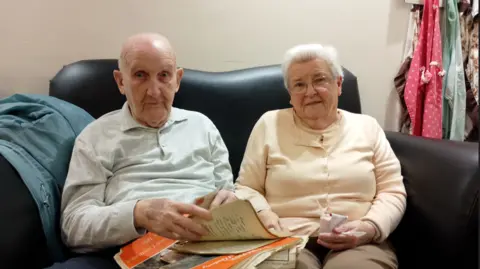 Ken Jack in grey shirt, holding orange vintage magazine, with Eleanor Jack beside him in cardigan, wearing glasses