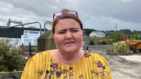 Joy Beard has brown hair and has a pair of pink glasses rested on her forehead
She is wearing a a multi-coloured floral pattern top. Ms Beard is standing outside to what appears to be a construction site. With a crane and heavy machinery visible in the background. 