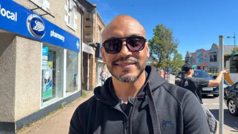 Jorge has a shaved head, is smiling and wearing sunglasses. He is standing outside in a street in Invergordon. There is a Boots pharmacy to the left of the picture. The sun is shining and the sky is blue.