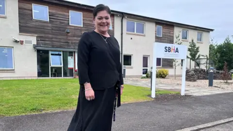 A woman with dark hair and a black dress stands outside an office building