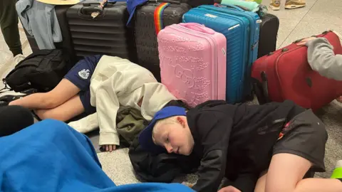 Jane Davis Lanchester EP Primary School pupils laying on the floor at Dubai airport while waiting for information after their Heathrow flight was cancelled. There are suitcase and bags around them.