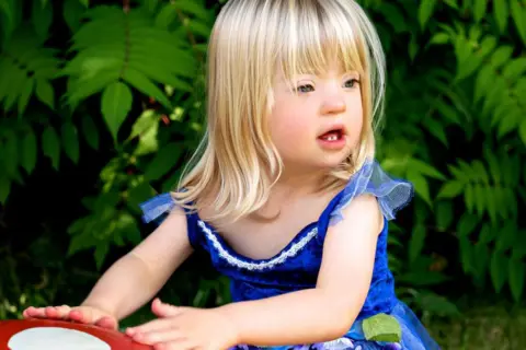 Five-year-old Martha. She has blond hair and wears a blue dress. She is playing in a garden and has her hands on what seems to be a red and white toadstool.