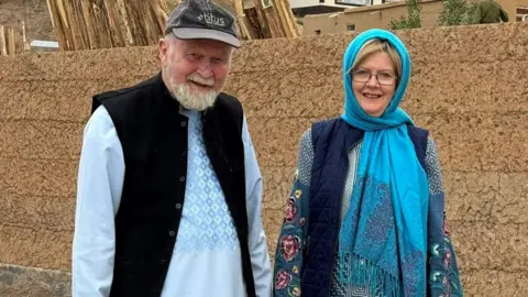 A man wearing a light blue robe and dark jacket with grey hair and a beard standing next to a woman in a blue dress with a turquoise scarf wrapped around her head  and wearing glasses in front of a brick wall