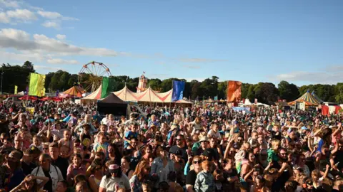 RICHARD WALPOLE A large crowd at an outdoor summer festival surrounded by tents and carnival rides.