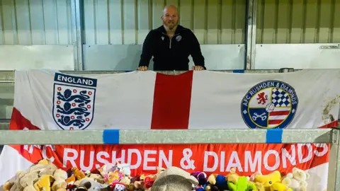 X/AFC Rushden & Diamonds Man standing on a football terrace, with an England flag spread out in front of him, at waist height, with AFC Rushden & Diamonds written in white capitals across the red horizontal stripe. A mound of soft toys can be seen at the bottom of the image.