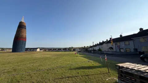 A very tall bonfire with a pyramid-shaped structure on top of it stands in the middle of a field in a housing estate. Around the outskirts of the field are rows of houses. 