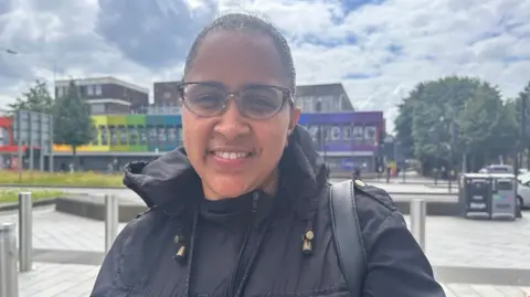 Vera Santos has short, wavy hair tied back, dark glasses and is a wearing a dark coat. She's also carrying a handbag. 