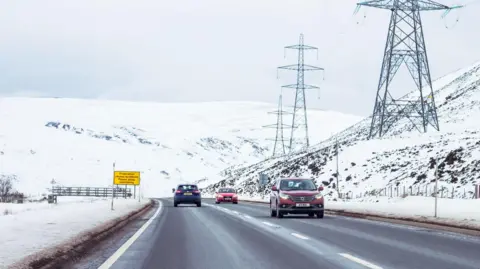 Getty Images Three cars travel along the A9 where it passes through the hilly Drumochter Pass. There is snow on the ground and, on the right hand side, large electricity pylons.