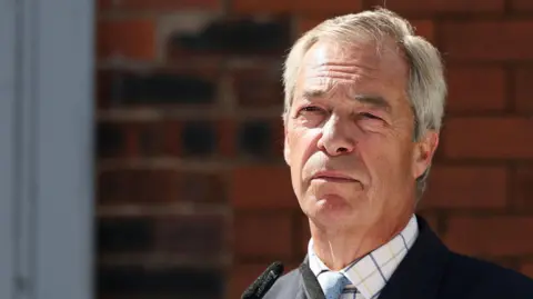 Nigel Farage standing outside in the sun. He has grey hair and is squinting, wearing a white shirt with a check pattern and a tie with the light blue of his party Reform. 