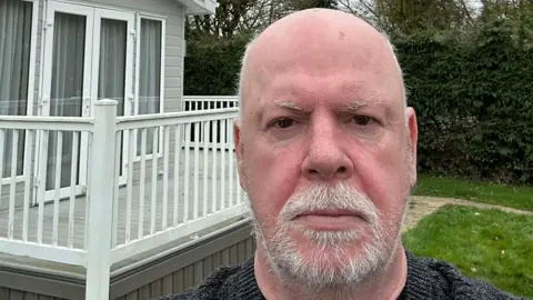 Supplied A head and shoulders selfie-style image of Gregg Dumbrell. He is standing in a caravan park in front of his lodge, which is located behind him to the left of the image.