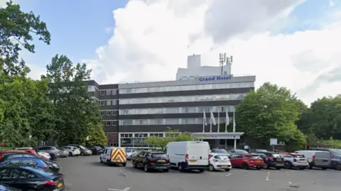 Google A picture of the Gosforth Park Hotel. It is a five-storey building with windows and grey bricks. Cars are parked outside, with trees on either side of the car park.