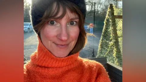 A selfie of a woman in an orange polo neck. She has long dark hair tucked into the neck of her hoodie and is wearing a black hat. She is standing in a car park and the sun is behind her.