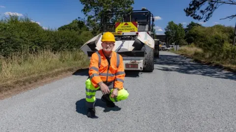 Leicestershire County Council A man in high-vis kneels in the road in front of a highway maintenance vehicle