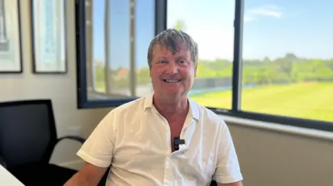 Jeff Evans, sat by a large window on a sunny day. He is wearing a white linen shirt, and has short dark grey hair.