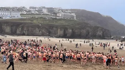 A group of many people on a beach, running towards the sea in swimwear.
