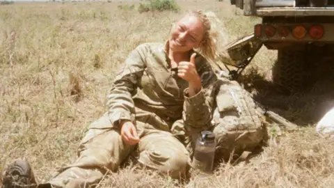 A 25-year-old girl with long, blonde hair tied in a ponytail smiles and gives a thumbs up as she sits in a field on a warm summer's day. She is wearing a camouflage Army uniform, and looks to have a light coating of paint or mud on her cheeks. Next to a water bottle, a camouflage backpack, and the rear of a truck-like vehicle.