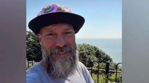 A screenshot of a selfie-style video of a man with a beard wearing a hat and a grey T-shirt. He is standing on the seafront and the sea is in the background. 