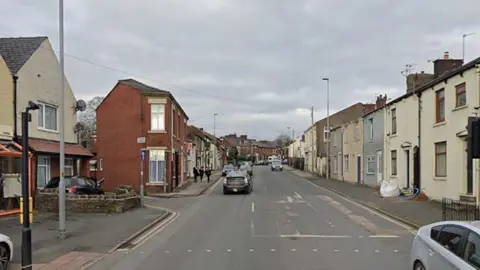 Google Street view image of Redlam, a three lane road that curves off to the right in the distance. There are red brick terraced shops and houses to the left and rendered terraced houses to the right