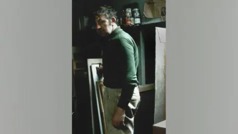 Gallerina A young Tom McGuinness in his loft space studio in Bishop Auckland. He is standing side on to the camera and is wearing a green top. He is standing in front of a number of canvases and tins of paints.