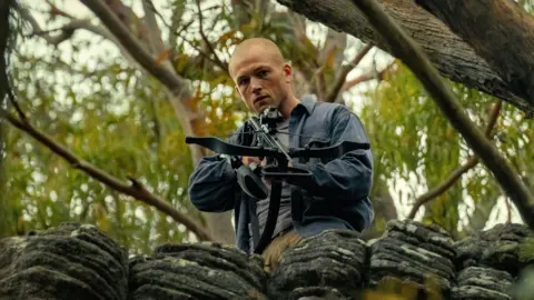Netflix Egerton is above the camera in a forest, aiming a crossbow. He wears a blur shirt over his grey t-shirt
