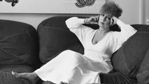 Parkin in a white dress lays on a sofa. She is looking at her camera. The photo is a black and white image.