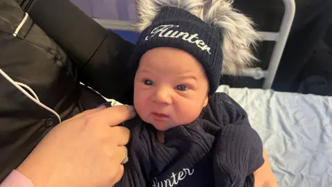A baby boy wearing a dark hat and top with the name Hunter embroidered into it