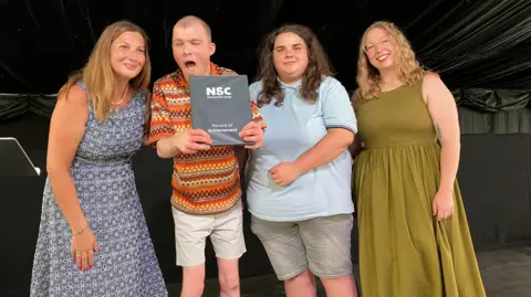 Four people stand on a stage posing for a photograph. One person holds a National Star College Record of Achievement certificate while the group stands together in front of a black backdrop.
