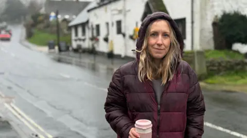 Local councillor Claire Critchison standing alongside a road, with her coat hood up while it rains
