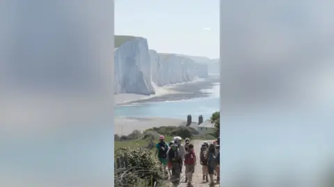 A group of people walk along a coastal path with white cliffs in the background