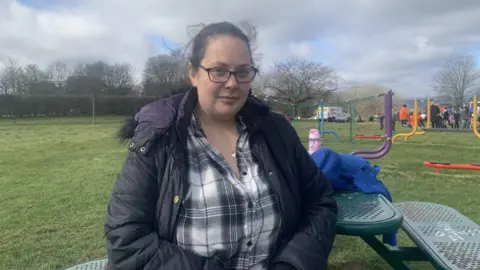 A woman has black hair tied back and glasses. She has her nose and chin pierced and is wearing a black hooded, padded coat and a black and white checked shirt. She is sitting at a table in a children's play area