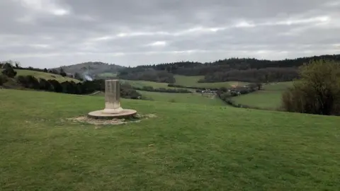 A view at Pewley Down in Guildford. In the middle of the image is a grey rectangular memorial stone on a pedestal. In the background there are fields and trees, including some woodland.