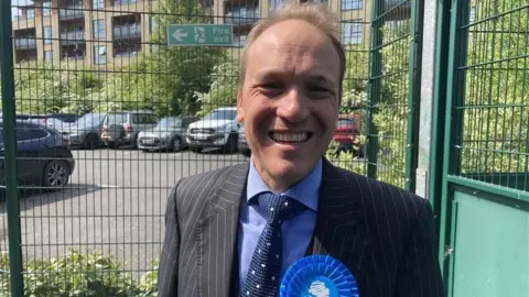 A man with short light brown hair smiles broadly at the camera, squinting in the sunlight. He is wearing a mid-blue shirt, a dark blue knitted tie and a grey pinstripe suit jacket.