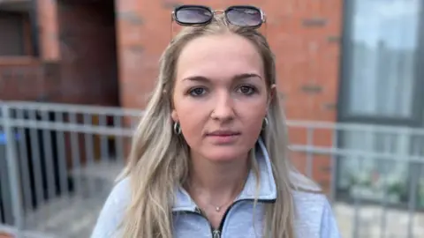 A woman with long blonde hair, wearing a grey jumper with sunglasses placed on top of her head, she is stood outside a red brick house with metal railings and is staring expressionless at the camera.