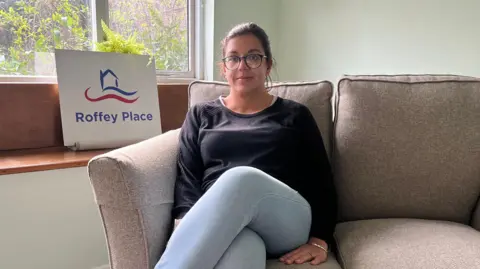 Alexis Green/BBC Tara Sims sitting on a break sofa. She is wearing glasses, blue jeans and a black jumper. She is looking directly at the camera, with a sign reading Roffey Place on it behind her. 