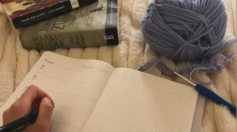 Supplied A partial hand, holding a pen, poised over an open page of an empty diary. In the top right is a ball of wool with a crochet needle, and two books in the top left.
