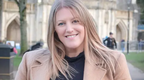 BBC A woman with long blond hair wearing a beige coat and a black top .