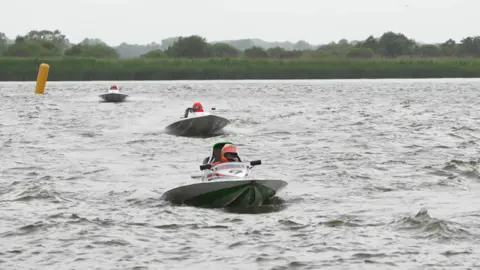 Three powerboats whizz towards the camera on a choppy river, with marsh and trees in the background. The three drivers wear orange helmets.