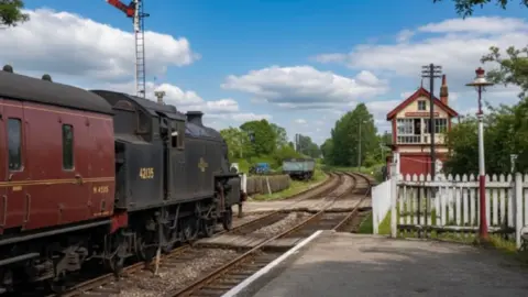 Edwin Williams The restored photo of New Longton and Hutton railway station in 1963 showing a train on the tracks to the left and the signal box building to the right behind a white picket fence. The train is now black with a deep red carriage, the signal box is white and red beside greenery and the sky is blue