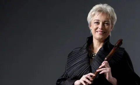 Malcolm Arnold Festival Michala Petri, holding a recorder, wearing a black top, with a necklace. She has short fair hair and is smiling to the camera.